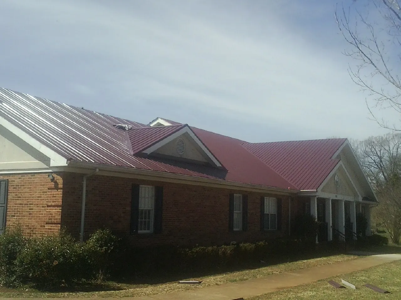 Red metal roof with gabled entries on a long brick building, from a Hail Damage Roof Repair job in Malta